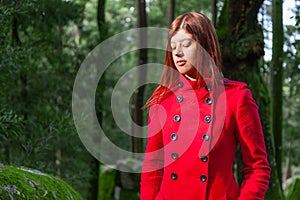 Young woman feeling sad walking alone on forest path wearing red long coat