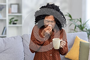 Young woman feeling unwell with cold or flu, sneezing into tissue, holding mug, at home on cozy sofa with cushions