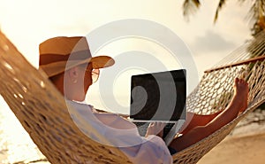 Young woman female freelancer using laptop while lying in hammock on the tropical beach at sunset photo