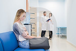 Young woman filling nervous sitting at hospital corridor waiting while two doctors talking on a backgroud