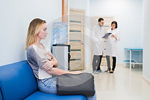 Young woman filling nervous sitting at hospital corridor waiting while two doctors talking on a backgroud