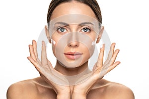 Young woman with flawless skin applying nourishing cream on her face cheek and fingers, looking at camera, studio white background