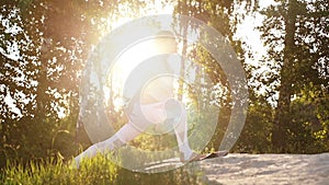 Young woman with flexible body exercising pose of warrior background of bright sunray and trees.