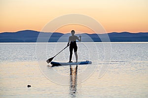 A young woman floats on a sapa on a mountain lake against the backdrop of sunset. Active recreation for a healthy lifestyle