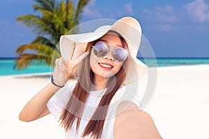 Young woman with a floppy sun hat and sunglasses takes a selfie in a tropical paradise photo