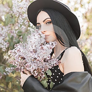 Young woman in flowered park rests in nature. Fashionable girl in elegant hat in trendy black dress and lilac spring flowers