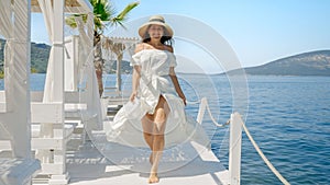 Young Woman in a Flowing Light Dress and Straw Hat Joyfully Runs along a Wooden Pier at the Beach, Embodying the Essence