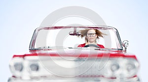 A young woman with fluttering hair rides in a red toy convertible, front view, on a light blue background