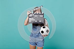 Young woman football fan with soccer ball, covering face with classic black film making clapperboard isolated on blue