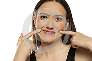 Young woman forcing her smile with her fingers on white background