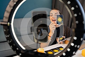 Young woman fortune teller recording video for blog using tarot cards, smartphone and ring lamp, predicts future online