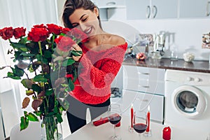 Young woman found red roses with candle, wine and gift box on kitchen. Happy girl smelling flowers. Women`s day