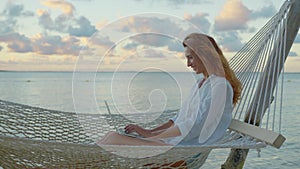 Young woman freelancer lying in a hammock with a laptop on the tropical beach at the sunset time on the background of