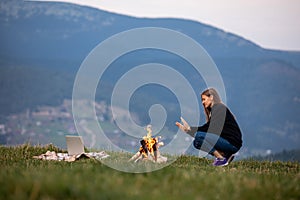 Young woman freelancer sits by the fire and warms his hands in the mountains in the evening. tourist girl resting