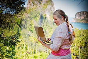 Young woman freelancer in white working with laptop on a top of the mountain with view on sea and tropical beach. Work and travel
