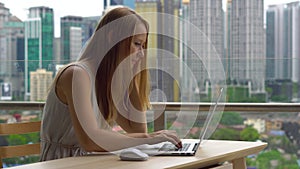 Young woman freelancer workes on her laptop at a balcony with a background of a city center full of skyscrapers. She