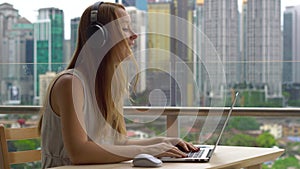 Young woman freelancer workes on her laptop at a balcony with a background of a city center full of skyscrapers. Remote