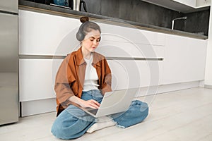 Young woman freelancer, working from home, student sitting with laptop on kitchen floor, wearing headphones, typing on