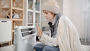 Young woman freezing in cold house warming at electric radiator. Concept of energy crisis, high bills, economy and saving money on