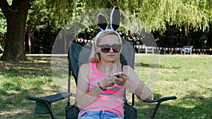 Young woman with funny rabbit ears hat sits in a chair and playing mobile in the park. Slow motion