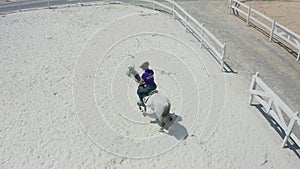 A young woman gallops on a white Arabian horse on a white sand court in an equestrian club. Aerial drone view.