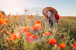 Young woman gathering poppies flowers walking in summer field enjoying landscape. Stylish happy girl wearing straw hat