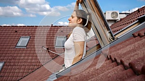 A young woman gazes out of an open attic window with a red tiled roof in the background and breathes fresh air