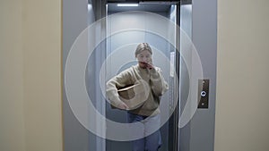 Young woman getting out of the elevator with cardboard box and talks on phone, moving in to new appartment