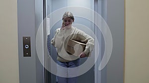 Young woman getting out of the elevator with cardboard box and talks on phone, moving in to new appartment