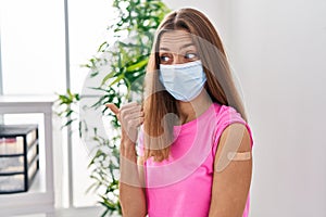 Young woman getting vaccine showing arm with band aid pointing thumb up to the side smiling happy with open mouth