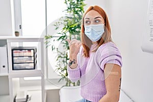 Young woman getting vaccine showing arm with band aid pointing thumb up to the side smiling happy with open mouth