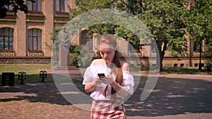 Young woman with ginger hair and bare shoulders typing on phone and walking in park near university building, greeting