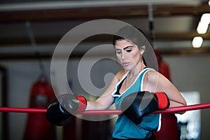 A young woman girl is hanging on the ropes of a boxing ring, resting, wearing boxing gloves.