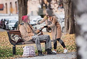 Young woman giving food to homeless beggar man sitting on a bench in city.