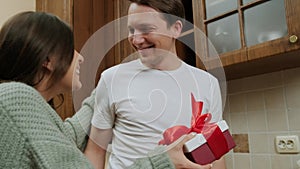 Young woman giving gift to her husband who prepares food in the kitchen.