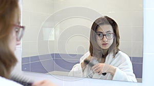 Young woman with glasses combing her hair in front of a mirror. A girl in a white bathrobe in front of a bathroom mirror