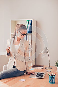 Young woman in glasses with cup of coffee sitting on table and typing on laptop