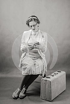 Young woman with glasses, dressed in retro style, sitting on a chair, with a suitcase at her feet, reading a book