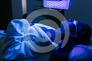 Young woman in glasses having blue LED light facial photodynamic therapy treatment in beauty salon. Female client having photo