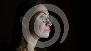 Young woman in glasses looks thoughtfully out the window from a dark room