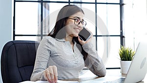 A young woman with glasses in the office is working on a laptop and talking on the phone