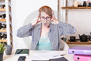 Young woman with glasses in shock holds her head with her hands over a stack of documents lying on the desk