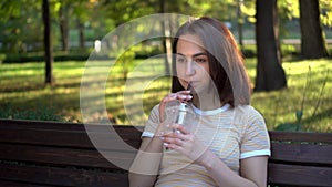 A young woman with glasses sits on a bench in the park and drinks cold coffee closeup. In the background there is a lake