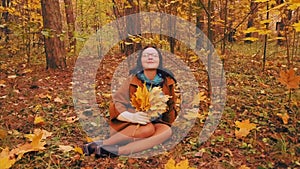 A young woman with glasses sits on the grass in the autumn park with maple leaves in her hands