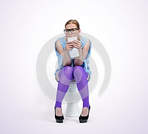 A young woman with glasses is sitting on the toilet holding toilet paper in her hands, on light background