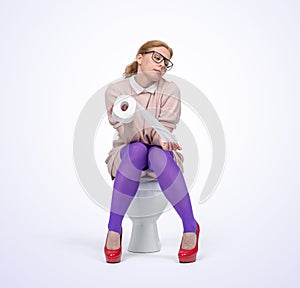 A young woman with glasses is sitting on the toilet holding toilet paper in her hands, on light background