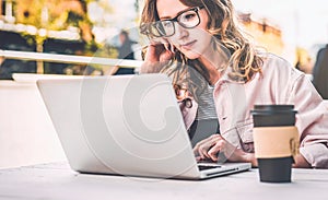 Front view.Young woman in glasses is sitting at table in street cafe and using laptop. Businesswoman working.