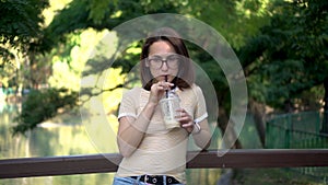 A young woman with glasses stands on a bridge in the park and drinks cold coffee closeup. In the background there is a