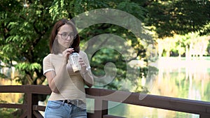 A young woman with glasses stands on a bridge in the park and drinks cold coffee closeup. In the background there is a