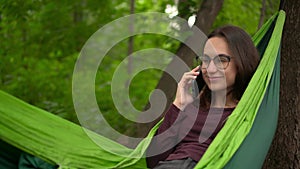 Young woman in glasses talking on the phone while lying in a hammock. A girl with glasses is resting in a hammock in